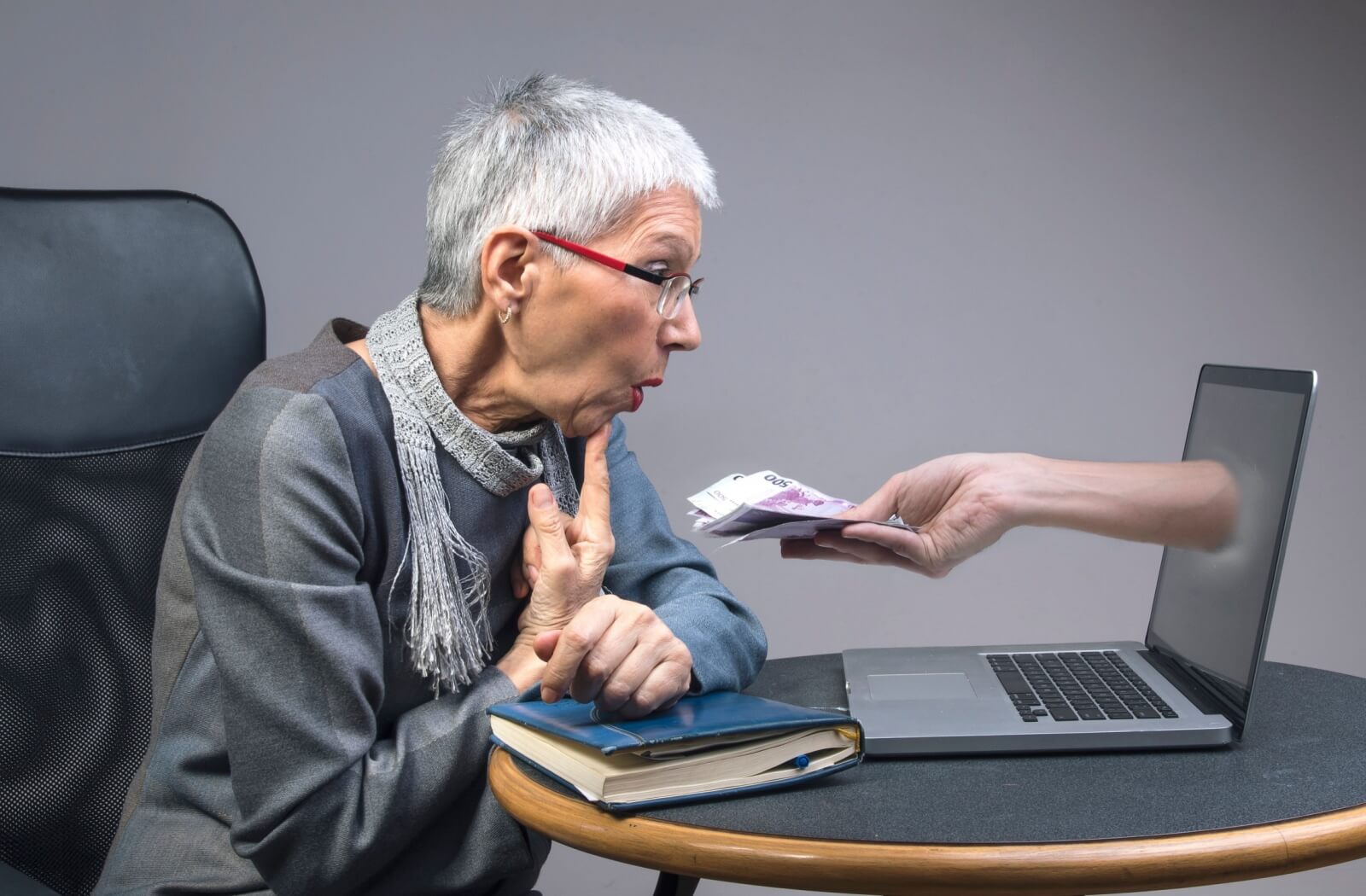 A senior gasps as a hand emerges from their laptop to offer a handful of cash, representing an online scam
