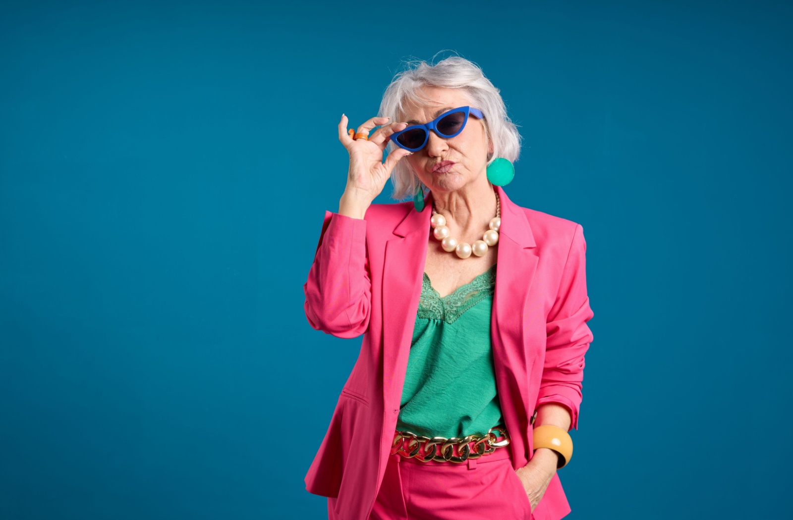 Against a blue backdrop, a stylish senior in a pink blazer and matching dress pants poses with sunglasses and chunky jewelry