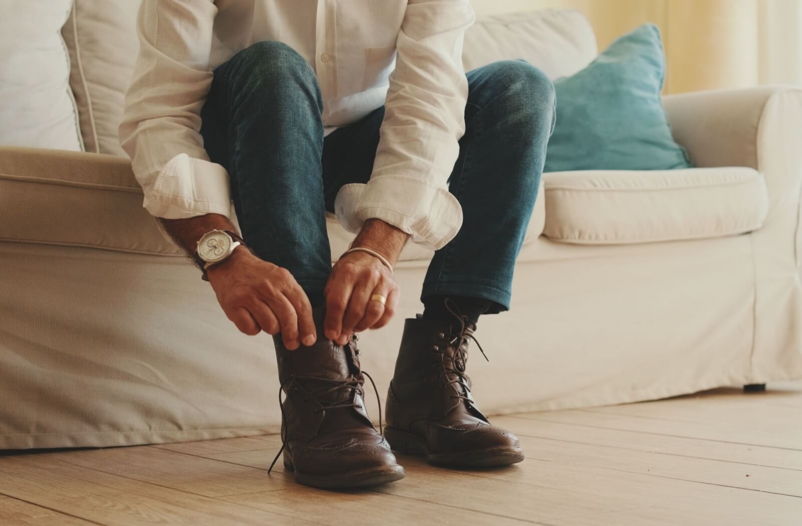An older adult in a white linen shirt carefully puts on a pair of stylish brown boots with great arch support