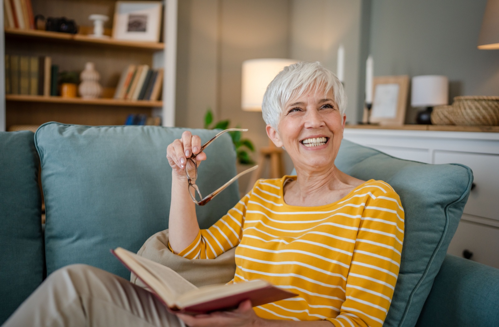 An older adult smiles as they relax on a blue couch while reading a book and holding their reading glasses