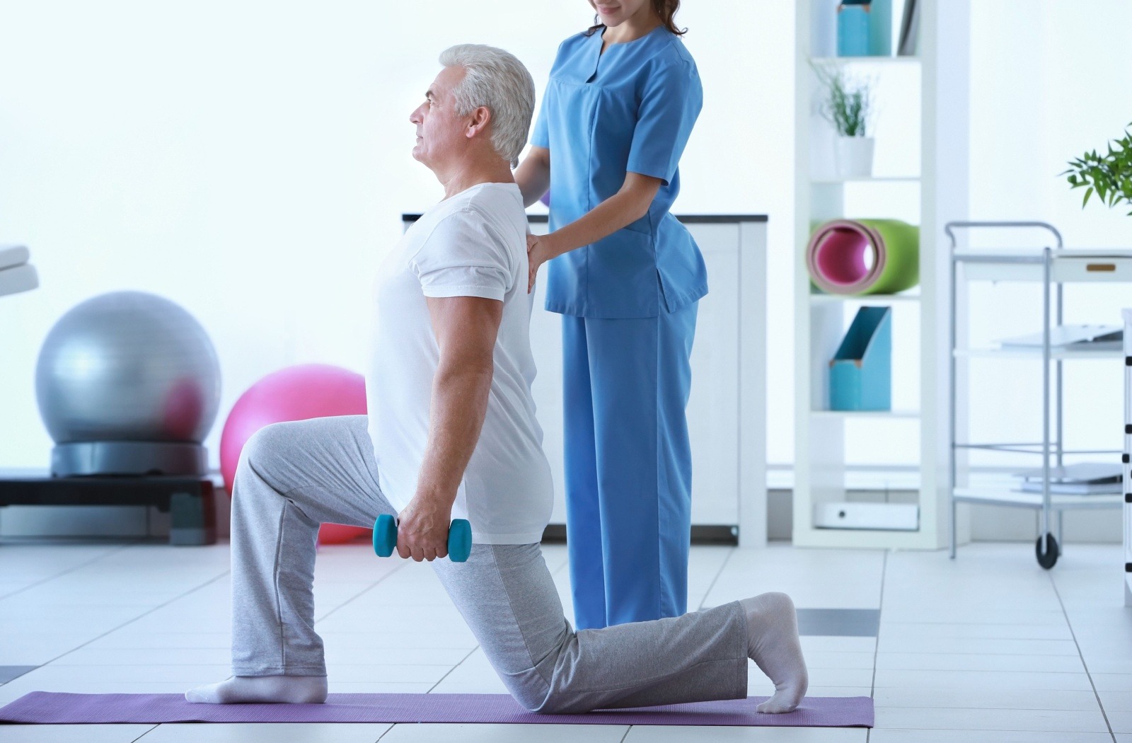 Under the guidance of a caregiver, a senior performs low-impact lunges with small dumbbells.
